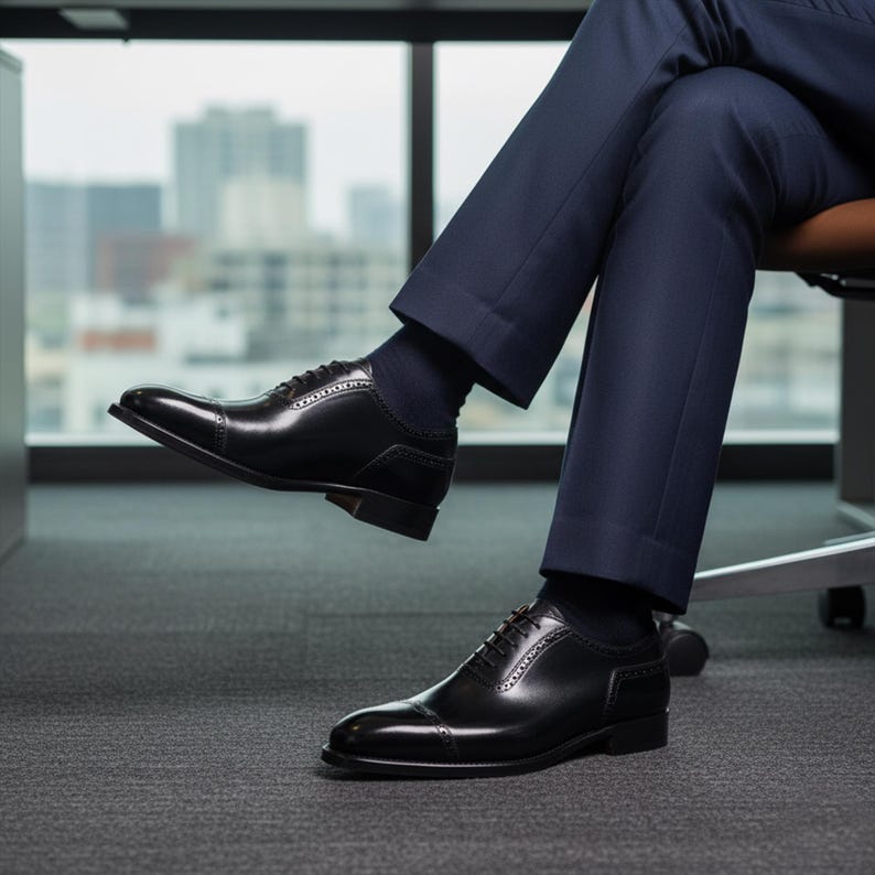 Puede incluir: Un par de zapatos oxford de cuero negro pulido con dise&ntilde;o de puntera y sutiles detalles brogue a lo largo de las costuras y los paneles de los ojales. Los zapatos se usan con pantalones de vestir azul marino oscuro y calcetines oscuros.