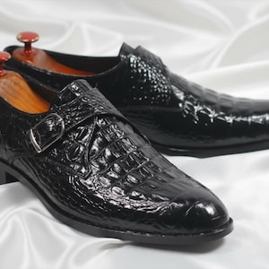 May include: A pair of black monk strap shoes featuring a glossy crocodile skin texture, a silver-toned metal buckle, and wooden shoe trees inserted inside each shoe to maintain their shape and structure.