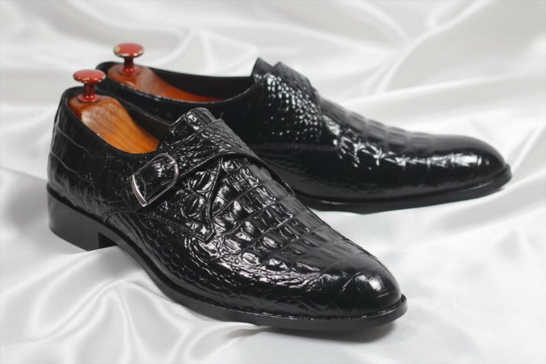 May include: A pair of black monk strap shoes featuring a glossy crocodile skin texture, a silver-toned metal buckle, and wooden shoe trees inserted inside each shoe to maintain their shape and structure.