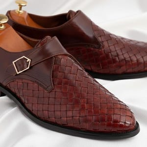 May include: A pair of dark brown leather shoes featuring a woven texture on the toe and side panels. Each shoe has a smooth leather strap across the top secured with a gold-toned metal buckle and a wooden shoe tree inserted inside the heel area.