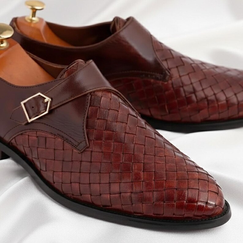 May include: A pair of dark brown leather shoes featuring a woven texture on the toe and side panels. Each shoe has a smooth leather strap across the top secured with a gold-toned metal buckle and a wooden shoe tree inserted inside the heel area.