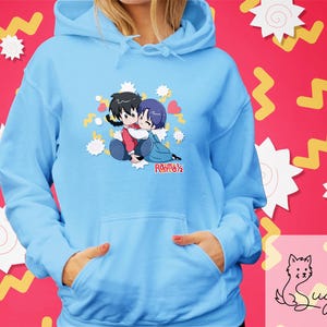 Può includere: Felpa con cappuccio azzurra con una grafica di due personaggi anime che si abbracciano, con il testo "Ranma ½" sotto. La felpa ha una tasca a marsupio e un cappuccio con coulisse. Lo sfondo è rosa con forme gialle e bianche.