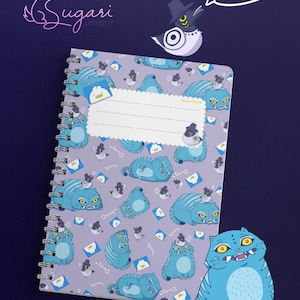 Puede incluir: Un cuaderno de espiral con un patrón repetido de gatos azules de dibujos animados con sombreros sobre un fondo lavanda. El cuaderno tiene una etiqueta en blanco y las palabras "3 Variations" en un bocadillo. El logotipo de Sugari Designs está en la esquina superior izquierda.