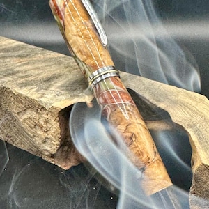 May include: A handcrafted pen with a wooden barrel featuring a natural, multi-toned wood grain pattern. The pen has silver-colored metal accents and a clip. The pen is displayed on a piece of wood, with wisps of smoke in the background.