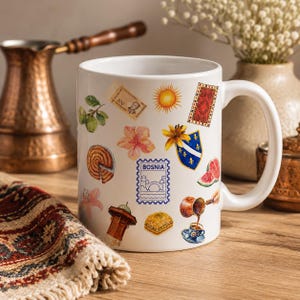 Op de afbeelding: Een witte keramische mok met een kleurrijke collage van illustraties, waaronder een Bosnische postzegel, een zon, een tapijt met patronen, een blauw schild met gele lelies, een stuk watermeloen, baklava, een koffiepot en een fontein.