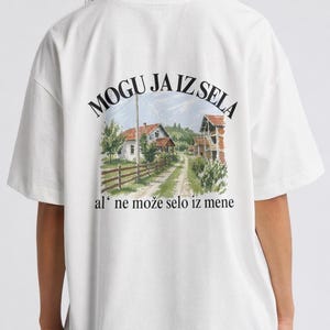 Könnte beinhalten: Weißes T-Shirt mit einer Landschaftsgrafik auf dem Rücken, die eine Straße zu Häusern zeigt, und dem Text "MOGU JA IZ SELA al' ne može selo iz mene."