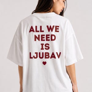 May include: A white short-sleeved t-shirt featuring the dark red text ALL WE NEED IS LJUBAV printed on the back, centred above a small dark red heart icon. The garment has a loose, relaxed fit with a crew neckline and short sleeves.