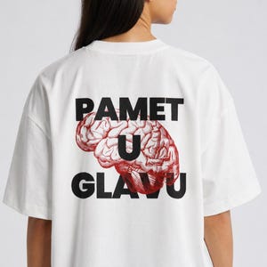 Op de afbeelding: Wit T-shirt met een grafisch ontwerp op de achterkant. Het ontwerp toont het woord "PAMET" boven een rode hersenillustratie, met het woord "U" in het midden en het woord "GLAVU" eronder.