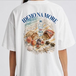 Könnte beinhalten: Weißes T-Shirt mit einem Strandpicknick-Motiv auf dem Rücken. Das Design zeigt eine Wasserflasche, Brot, Pfirsiche, Feigen, Snacks, Sonnenbrillen und den blauen Text "IDEMO NA MORE".