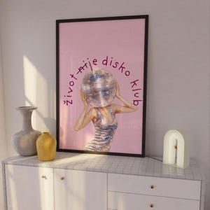 May include: Framed art print with a pink background, depicting a woman with a disco ball head. The text "život nije disko klub" is written around the disco ball. The print is displayed on a white cabinet with decorative vases and a lamp.
