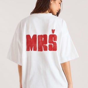 Könnte beinhalten: Weißes übergroßes T-Shirt mit dem Wort "MRS" in Rot auf dem Rücken gedruckt. Das Shirt hat kurze Ärmel und eine lockere Passform. Der Text ist in einer fetten, serifenlosen Schriftart.