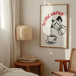May include: A framed art print with a black and white coffee illustration and the red text "Jutrić Kafica". The print is in a wooden frame, hanging on a white wall. A woven lamp and side table are also visible.