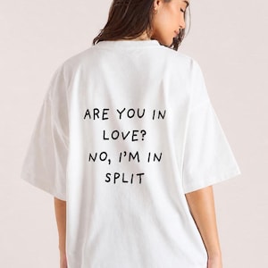 Könnte beinhalten: Weißes T-Shirt mit dem Text "ARE YOU IN LOVE? NO, I'M IN SPLIT" in Schwarz. Das Shirt hat kurze Ärmel und eine lockere Passform.