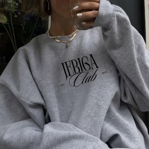 Könnte beinhalten: Ein graues Sweatshirt mit dem schwarzen Schriftzug "JERIGA Club" in einem stilisierten Schriftstil. Das Sweatshirt hat auch den Text "EST. 2025". Der Stil ist lässig.