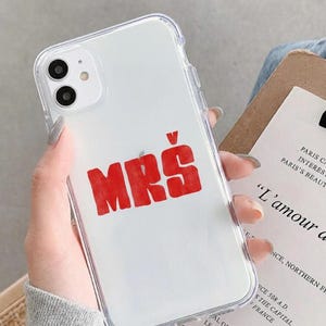 Puede incluir: Funda transparente para teléfono con la palabra "MRS" en letras rojas sobre un fondo blanco. La funda está colocada en un teléfono inteligente blanco con una cámara de doble lente. El teléfono se sostiene cerca de un papel con texto.
