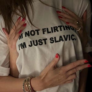 Może przedstawiać: Biała koszulka z napisem "I'M NOT FLIRTING, I'M JUST SLAVIC" w kolorze czarnym. Koszulka jest obejmowana przez ramiona z czerwonym lakierem do paznokci i złotą biżuterią. Na lewym nadgarstku widoczny jest stos bransoletek.