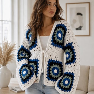 Op de afbeelding: Een gehaakt vest met lange mouwen en een granny square patroon in wit, zwart, blauw en saliegroen. Het kledingstuk heeft een V-hals, wijde mouwen en een striksluiting aan de voorzijde van wit garen, gedragen over een effen wit hemd.
