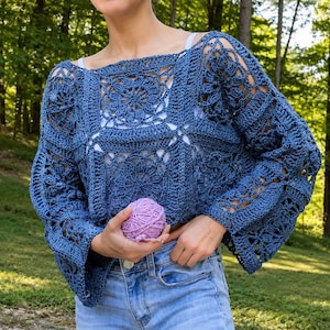 Könnte beinhalten: Ein blaues, langärmeliges Häkeloberteil mit einem quadratischen Lochmuster und Blumenmotiven. Das Kleidungsstück hat einen weiten Ausschnitt und lockere Ärmel, wird über einem weißen Unterhemd zu einer hellblauen Jeans getragen und hält ein Garnknäuel.
