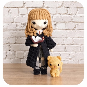 May include: A crocheted doll with light brown yarn hair, wearing a black robe over a grey skirt, white shirt, and striped tie, holding an open book. Beside the doll stands a small, light orange crocheted cat with pointed ears and black eyes.