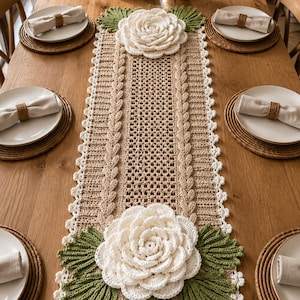 Può includere: Un runner da tavola rettangolare all'uncinetto color beige con un motivo centrale a trama aperta, fiancheggiato da bordi intrecciati e rifinito con grandi fiori tridimensionali all'uncinetto bianchi e foglie verdi alle estremità.