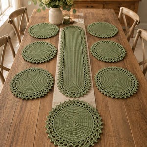 Op de afbeelding: Een set van zeven saliegroene gehaakte tafelaccessoires bestaande uit één lange ovale tafelloper en zes bijpassende ronde placemats. Elk stuk heeft een dicht gehaakt midden en een decoratieve, opengewerkte rand met schulpen.