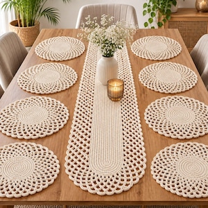 May include: A set of six round cream-coloured crocheted placemats and one matching long rectangular table runner with a scalloped edge design. The items feature a dense centre weave surrounded by an openwork pattern made of thick cord.