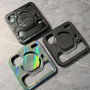 Puede incluir: Tres fundas cuadradas para un dispositivo, cada una con un diseño diferente. Una es negra con un patrón de vetas de madera, otra es negra con una superficie texturizada y la tercera es iridiscente con un brillo de arcoíris. Las fundas tienen recortes para botones y una pantalla.