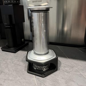 Puede incluir: Una cafetera AeroPress, plateada y negra, con el nombre de la marca en la parte superior. La cámara cilíndrica tiene marcas de medición. A la izquierda hay un molinillo negro. La cafetera está sobre una superficie gris.