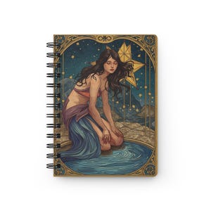Può includere: Quaderno a spirale con un'illustrazione celeste. L'opera raffigura una donna inginocchiata vicino all'acqua, con una stella e un cielo stellato sullo sfondo. La copertina ha un bordo color oro con un design ornato. Il quaderno è bianco.
