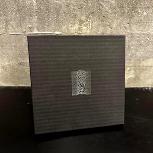 May include: A dark gray box with a textured surface. The box features a white graphic of a wave-like pattern in the center. The box is sitting on a black surface against a gray concrete wall.