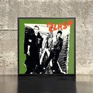 May include: Framed album cover featuring The Clash band members. The album art has a green background with a torn paper effect. The band's name, "The Clash," is in red. The band members are in black and white.