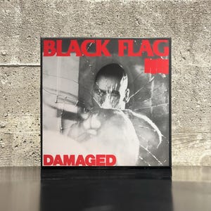 May include: A framed album cover for the band Black Flag. The album cover is black and white with a man's face in the center, and the words "BLACK FLAG" and "DAMAGED" in red block letters.