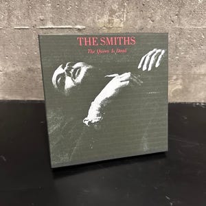 May include: A square album cover for The Smiths' "The Queen Is Dead" album. The cover features the band's name in pink text at the top, with the album title below. A black and white image of a person is in the center.