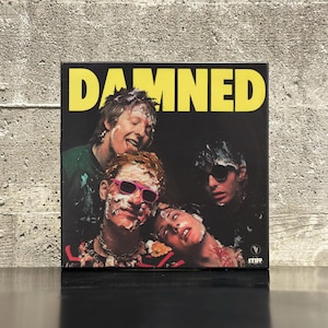 May include: A square album cover featuring the band The Damned, with the word "DAMNED" in large yellow letters. The image shows four band members covered in what appears to be cake or frosting. The album cover has the Stiff Records logo.