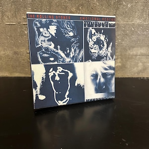 May include: A CD case featuring the album "Emotional Rescue" by The Rolling Stones. The cover art is a collage of blue and white images, including faces and abstract shapes. The band's name and album title are printed in red and white.