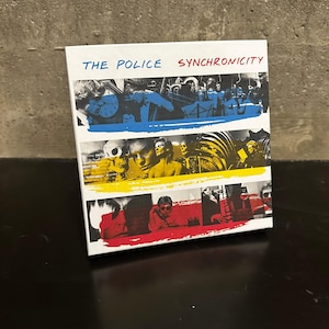 May include: A CD case for The Police's album "Synchronicity." The cover features a design with three horizontal bands in blue, yellow, and red, each with a different image of the band members. The band's name and album title are in white text.