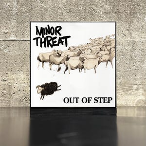 May include: A framed print featuring the band name "MINOR THREAT" above an illustration of sheep. The artwork includes a black sheep jumping away from the flock, with the words "OUT OF STEP" below the image. The print is displayed on a reflective surface.