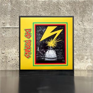 May include: A framed print with a yellow background and the words "BAD BRAINS" in green and red. The artwork features a black and white illustration of a building with a lightning bolt and starburst design.