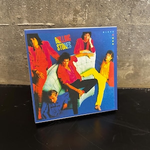 May include: Album cover for The Rolling Stones' "Dirty Work" album. The cover features a group of people in vibrant outfits against a blue backdrop. The album title is in a colorful, stylized font.
