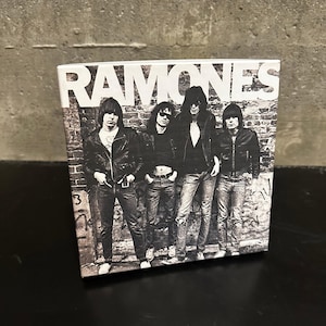 May include: A black and white image of the band The Ramones. The band members are standing in front of a brick wall. The word "RAMONES" is printed in large white letters at the top of the image.