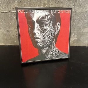 May include: A CD case featuring the Rolling Stones' album "Tattoo You." The cover art displays a black and white portrait with intricate tattoo-like designs on a red background. The album title is printed at the top.