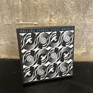 May include: A square box featuring a repeating black and white graphic design with a spiral and gear motif. The top of the box has the text "ROLLING STONES STEEL WHEELS" in blue and white.