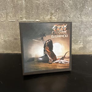 May include: A square, wooden box featuring album art. The artwork depicts a person in a seated position with arms raised, a cross, and the text "Ozzy Osbourne Blizzard of Ozz". The box is on a black surface.