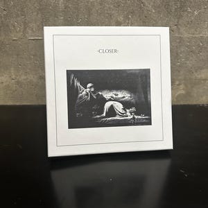 May include: A white square book with the word "CLOSER" printed at the top. The cover features a black and white image of a person lying down with another person sitting nearby. The book is on a black surface.