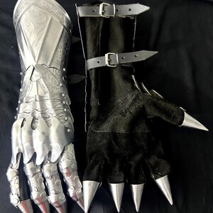 May include: A pair of medieval-style gauntlets. One is silver metal with ornate detailing, the other is black with silver claw-like tips. The black gauntlet has leather straps with buckles. The image includes the text "Battle Costume".