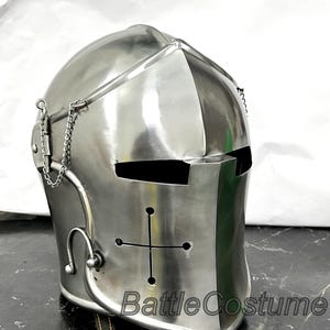 Puede incluir: Un casco medieval plateado con un diseño de cruz y el texto "Battle Costume". El casco tiene una visera con ranuras para los ojos y un detalle de cadena. El casco es de metal.