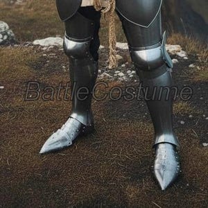 K&ouml;nnte beinhalten: Nahaufnahme einer polierten Beinr&uuml;stung aus Stahl, Teil eines mittelalterlichen Ritterkost&uuml;ms. Die R&uuml;stung umfasst Beinschienen, Kniekappen und Sabatons. Der Text "Battle Costume" ist auf dem Bild sichtbar.