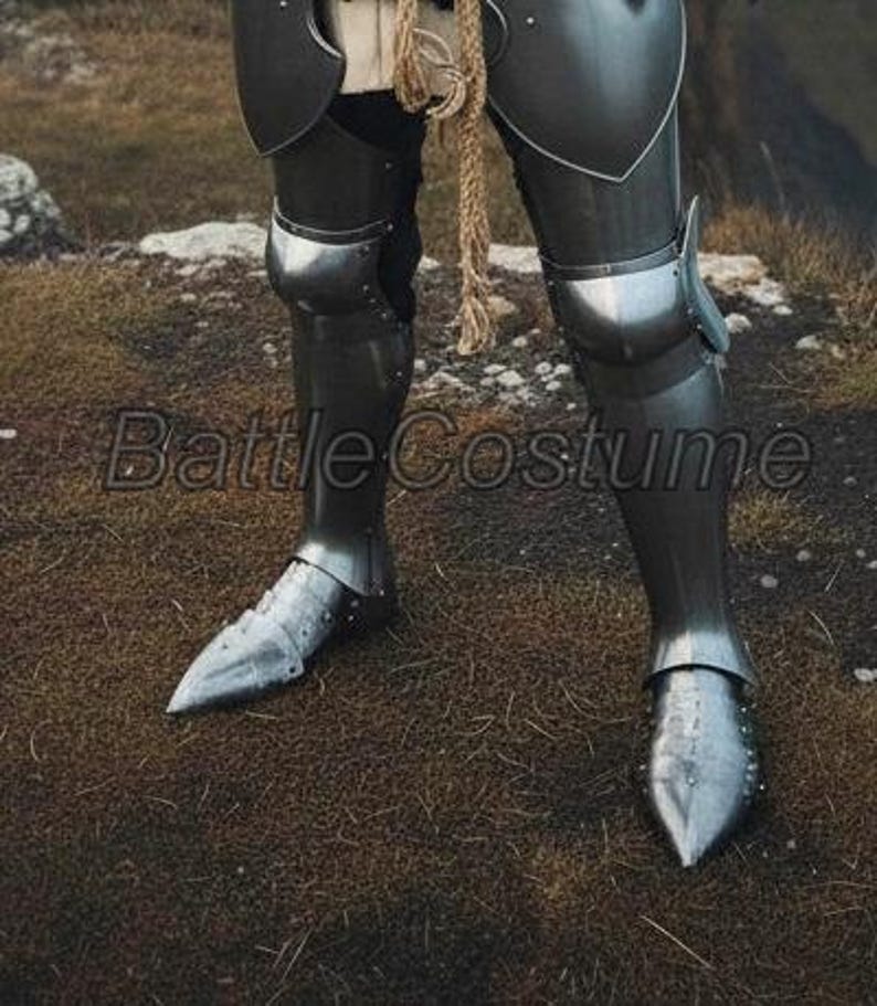 K&ouml;nnte beinhalten: Nahaufnahme einer polierten Beinr&uuml;stung aus Stahl, Teil eines mittelalterlichen Ritterkost&uuml;ms. Die R&uuml;stung umfasst Beinschienen, Kniekappen und Sabatons. Der Text "Battle Costume" ist auf dem Bild sichtbar.