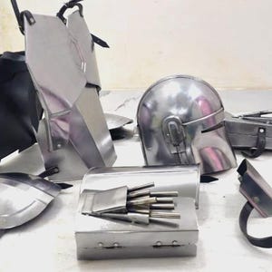 May include: A collection of silver-colored metal armor pieces, including a helmet, chest plates, and arm guards. The armor appears to be new and unused, with straps and hinges visible for assembly. The pieces are arranged on a light-colored surface.