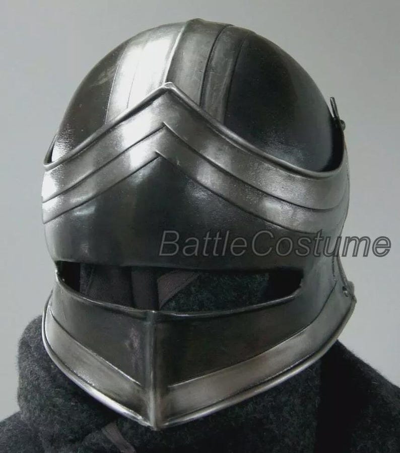 K&ouml;nnte beinhalten: Ein schwarzer mittelalterlicher Helm mit Visier und abgerundeter Oberseite. Der Helm hat silberfarbene Verzierungen und der Text "BattleCostume" ist auf der Seite sichtbar. Der Helm ist f&uuml;r Kost&uuml;me oder Cosplay konzipiert.